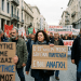 Woman holding sign reading “Ο 13ος ΚΑΙ Ο 14ος ΜΙΣΘΟΣ ΔΕΝ ΕΙΝΑΙ ΠΟΛΥΤΕΛΕΙΑ. ΕΙΝΑΙ ΑΝΑΓΚΗ.”; banner reads “ΔΙΚΑΙΩΣΗ ΣΤΟΥΣ ΕΡΓΑΖΟΜΕΝΟΥΣ.”