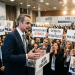 Greek political speaker at HELEXPO surrounded by attendees holding protest signs